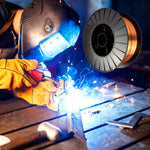 Arame para solda Mig 0,8 mm em uso com gás nobre, rolo de 15kg. Soldador utilizando o arame em processo de soldagem, com faíscas e brilho intenso, mostrando a alta performance e qualidade do produto para soldas precisas e resistentes em aço carbono e outras ligas metálicas.