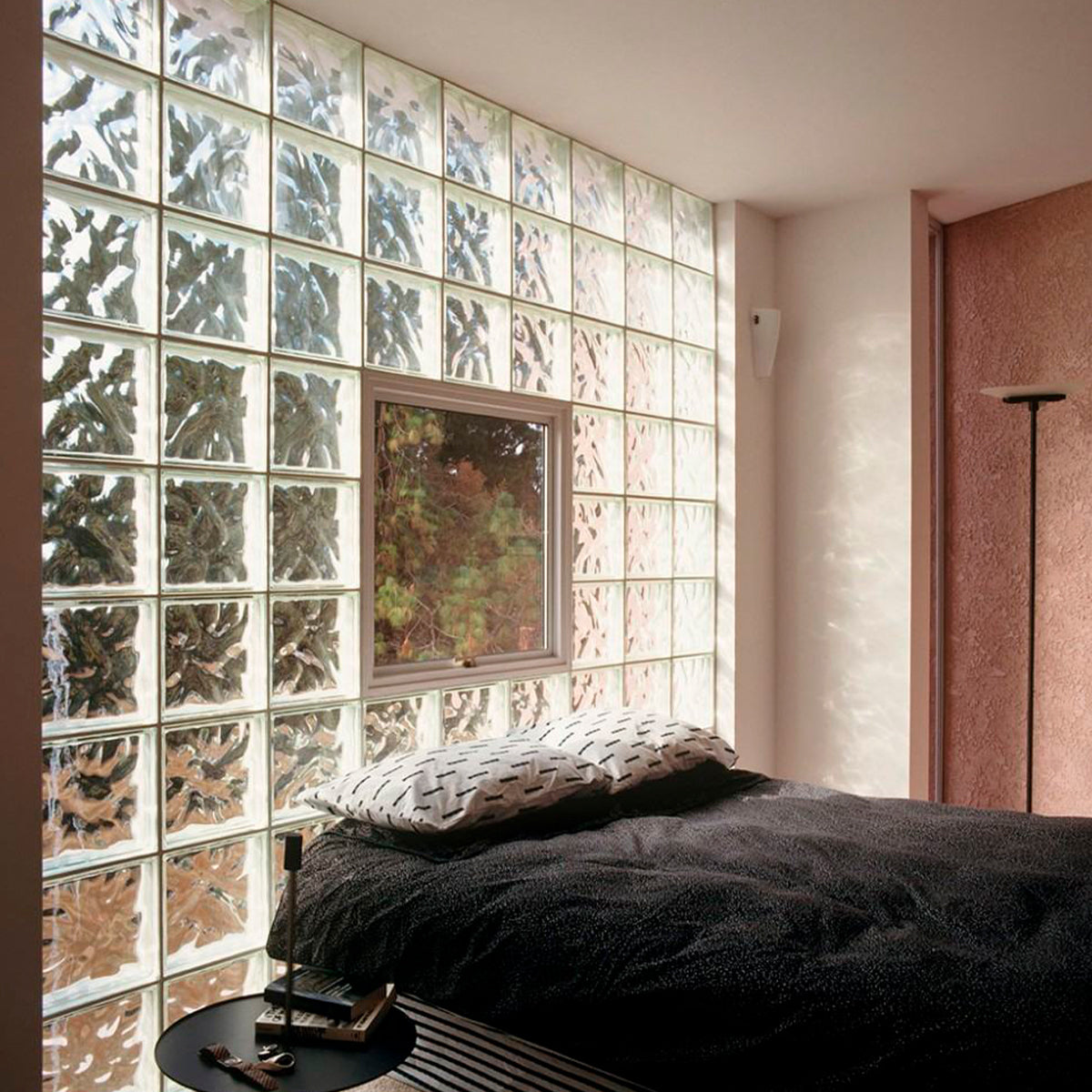 Quarto moderno com parede de blocos de vidro transparente Tradestar 19x19x8, permitindo a passagem de luz natural, com cama, travesseiros e janela com vista para área verde, ideal para divisórias e decoração de ambientes.