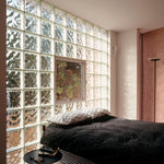 Quarto moderno com parede de blocos de vidro transparente Tradestar 19x19x8, permitindo a passagem de luz natural, com cama, travesseiros e janela com vista para área verde, ideal para divisórias e decoração de ambientes.